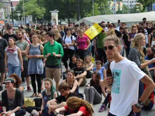 Ein Mann steht vor einer unüberschaubaren Menge von Zuschauer/innen und hält ein Getränk in der Hand. In seinen am Hinterkopf zusammengebundenen Haaren steckt ein Fähnchen, auf dem "Ja zu Flüchtlingsschutz" aufgedruckt ist.