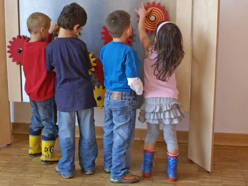 Drei Jungen und ein M&auml;dchen probieren bunt angemalte Holzzahnr&auml;der aus, die an einer Ausstellungswand verschraubt sind.
