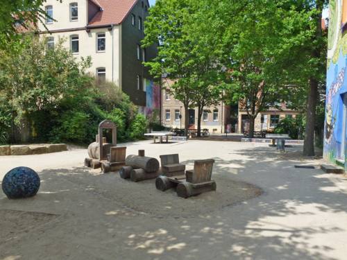 Auf dem Spielplatz in der Albertstraße sind unter Bäumen zwei Tischtennis-Platten zu sehen sowie eine Holzeisenbahn.