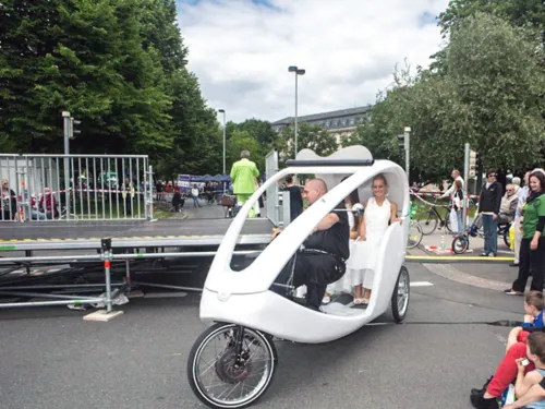 Zwei Frauen lassen sich in einer Fahrrad-Rikscha fahren, Passanten schauen interessiert zu