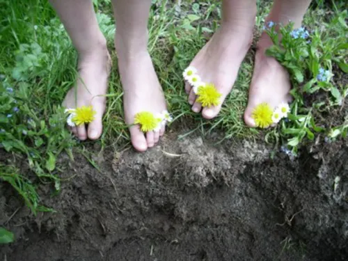Blüten von Löwenzahn und Gänseblümchen schmücken Füße, die auf Gras und schwarzer Erde stehen.