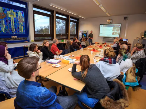 Schülerinnen, Schüler und Lehrkräfte sitzen um einen langen Tisch und hören einem Vortrag zu