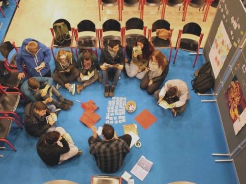 Schülerinnen und Schüler sitzen im Kreis auf den Boden neben Stellwänden und arbeiten mit Karten