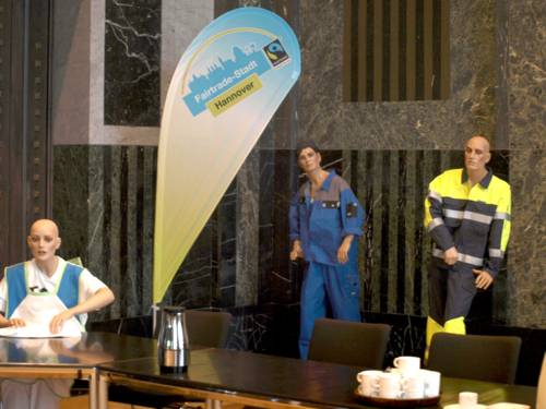 Drei Schaufensterpuppen sind mit Arbeitskleidung an und hinter einem Tisch zu sehen, daneben eine Fahne mit der Aufschrift "Fairtrade-Stadt Hannover"