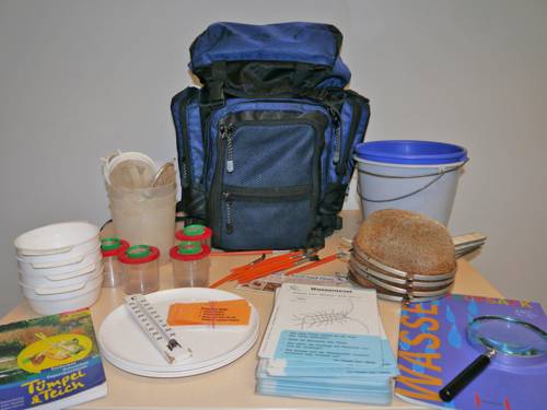 Ein blauer Rucksack steht zusammen mit vielen Utensilien, die man zur Untersuchung von Wasserproben benötigt, auf einem Tisch: Becher, Siebe, Bestimmungskarten, Eimer, Schalen, Broschüren