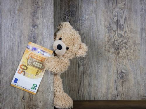 Ein Teddybär lugt hinter einer Holzwand hervor mit einem 50-Euro-Schein in der Hand.