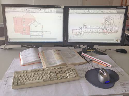 Zwei Monitore mit technischen Zeichnungen von Gebäuden und ein Schreibtisch, auf dem ein großformatiger Bauplan, Bücher, Stift und Lineal liegen