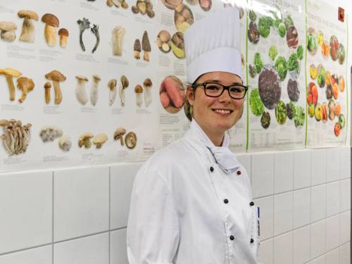 Eine junge Frau in weißer Koch-Berufsbekleidung und Mütze steht vor einer gefliesten Wand, die durch Poster von Pilzen und Gemüsesorten aufgelockert wird. 