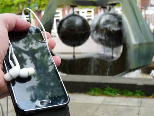 Jemand hält ein Smartphone mit Kopfhörern vor einem Brunnen in der Hand.