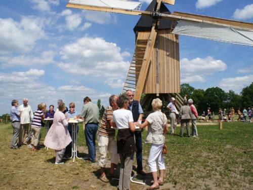 Menschen an Tischen im Freien vor einer wiederaufgebauten Windmühle.