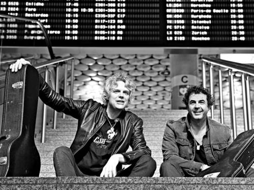 Zwei Männer mit Gitarrenkoffern sitzen auf einer Treppe, im Hintergrund eine Flughafen-Anzeigetafel.