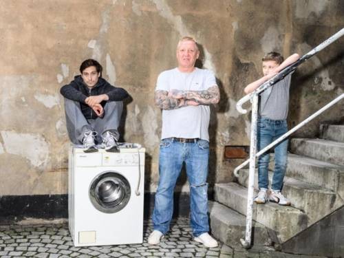 Ein Mann sitzt auf einer Waschmaschine. Daneben steht ein weiterer Mann und ein Junge auf einer Treppe. 