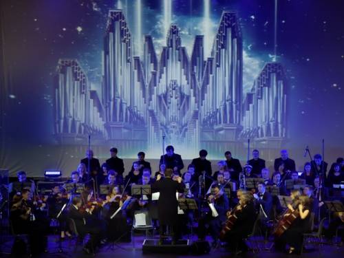 Ein großes Orchester ist zu sehen. Der Dirigent steht in der Mitte mit erhobenen Armen.