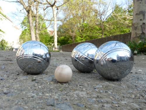 Drei große und eine kleine Metallkugel auf einer Sandfläche vor einem Park.