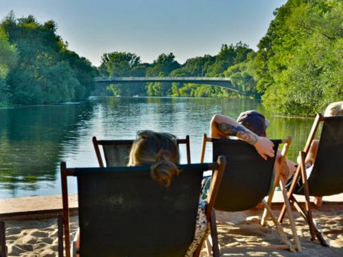 Zwei Personen in Sonnestühlen blicken auf eine Brücke über einen Fluss.