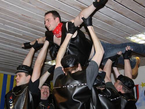 Männer mit Lederwesten und -mützen heben einen Mann in die Höhe.