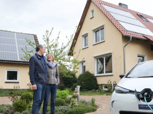 Frau und Mann stehen Arm in Arm vor einem Haus, im Vordergrund wird ein Elektroauto geladen.