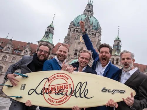 F&uuml;nf M&auml;nner halten freudestrahlend ein Surfbrett hoch, das die Aufschrift "Leinewelle genehmigt" tr&auml;gt