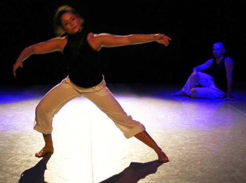 Eine dunkelhaarige Tänzerin bewegt sich mit weit ausgestreckten Armen, im Hintergrund sitzt ein Tänzer auf dem Boden und beobachtet sie.