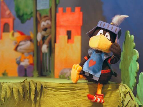 Szene aus einem Stück: Ein Rabe im Ritterkostüm sitzt vor einer Burg.