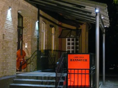 Ein Hauseingang mit überdachter Treppe und einem Schild mit der Aufschrift Jazz Club Hannover, in der geöffneten Tür steht ein Kontrabass.