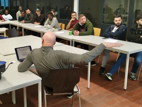 In einem Raum sitzen verschiedene Personen an Tischen, vor ihnen sitzt ein Mann, der einen Arm hebt