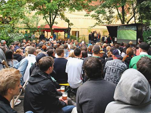 Viele Menschen in einem Biergarten vor einer Leinwand, auf der ein Fußballspiel gezeigt wird.