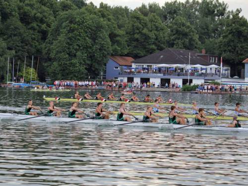 Foto: Voll besetzte Ruderboote auf dem Maschsee, dahinter am Ufer Zuschauer und das Gebäude des Ruder-Clubs mit Terasse.