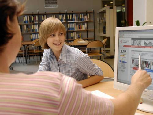 Eine Frau, die nur von hinten zu sehen ist, zeigt einer jungen Erwachsenen, die ihr gegenüber sitzt, etwas auf dem Computerbildschirm.