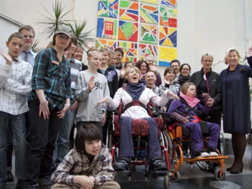 Mehrere Schüler einer Förderschule, zwei von ihnen im Rollstul, die vor einem selbstgemalten Kunstwerk stehen und fröhlich in die Kamera winken.
