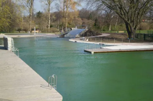 Überblick über das Naturbecken mit seiner hölzernen Beckenumrandung, im Hintergrund Rutsche und Sprungfelsen sowie zweites Becken und Liegewiese