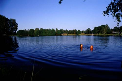 Ansicht des Sees, in dem zwei Menschen schwimmen. Im Hintergrund befinden sich viele Bäume.