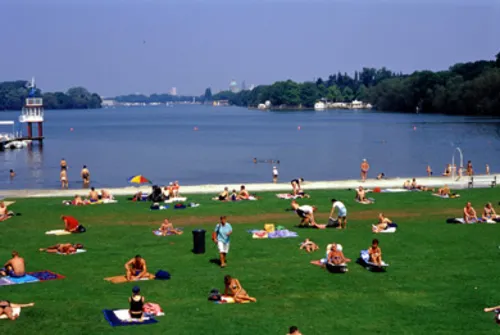 im Vordergrund liegen und sitzen Menschen in Badebekleidung auf Badetüchern auf Rasen, im Hintergrund ist ein dünner Streifen Strand und der Maschsee