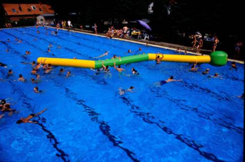 Blick von oben in ein Schwimmbecken, in dem Schwimmbecken befindet sich eine riesige aufgeblasene Raupe aus grünem und gelben Kunststoff, Schwimmer spielen damit und versuchen auf die Raupe zu klettern