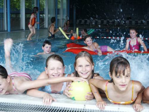 Vier Mädchen mit gelben Ball am Beckenrand, im Hintergrund im Wasser tobende Kinder