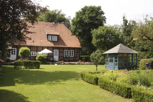 Gepflegtes, parkähnliches Grundstück mit Rasen und Hecken, im Hintergrund ein rotes Fachwerkhaus im Sonnenschein