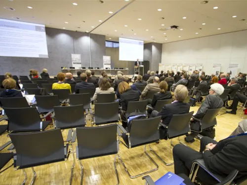 Kinobestuhlung in einem Sitzungsraum mit einigen nicht besetzten Plätzen. Vorne befinden sich zwei Leinwände, ein Podium und ein Pult, an dem gerade ein Redner spricht.
