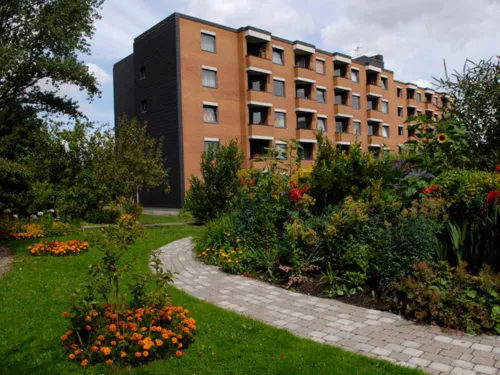Eine Wohnanlage, zu der ein kleiner gepflasterter Weg führt. Entlag des Weges sind viele schöne Blumen und insgesamt gibt es vor dem Haus viel Grün.