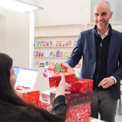 Eine Frau sitzt an einem Tisch, auf dem gestapelte rot eingepackte Pakete liegen. Ihr gegen&uuml;ber steht ein Mann, der ein weiteres P&auml;ckchen &uuml;berreicht.