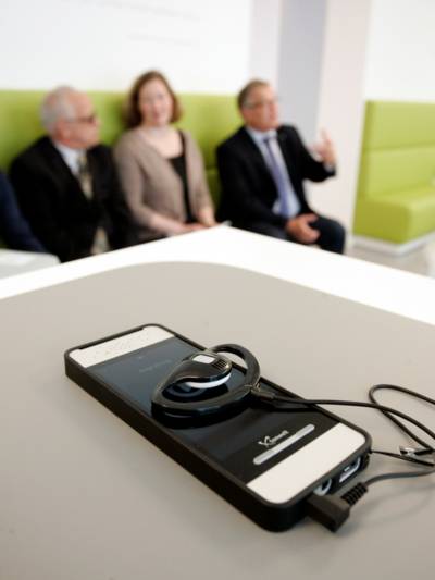 Ein Gerät, ähnlich einem Smartphone, mit einem Kopfhörer liegt auf einem Tisch. Im Hintergrund sind drei Personen zu sehen.