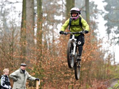 Ein Mointainbiker trägt eine hellgrüne Jacke und einen rosafarbenen Helm. Er fliegt gerade samt Fahrrad nach einem Sprung über dem Boden. Am linken Rand steht ein älterer Herr mit einem Jungen, beide schauen zu.