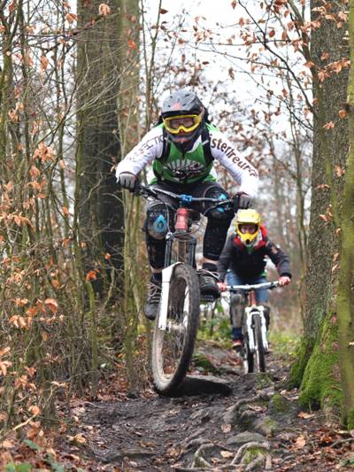 Zwei Mountainbiker tragen Schutzkleidung und fahren hintereinander über einen unebenen Weg mit zahlreichen Baumwurzeln.