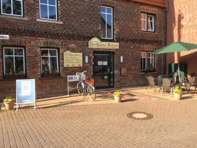 Ein rotes Backsteingeb&auml;ude, davor ein Werbeaufsteller, ein Fahrrad und eine Sitzm&ouml;glichkeit aus Gartenst&uuml;hlen, Tischen und einem Sonnenschirm