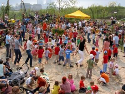 Zahlreiche Kinder und Erwachsene halten sich an den Händen und tanzen auf Sand um einen Maibaum herum