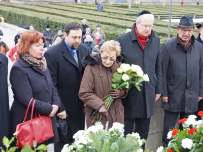 Zwei Frauen und drei M&auml;nner vor einem Mahnmal mit Blumen.