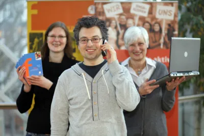 Ein junger Mann l&auml;chelt mit einem Telefon am Ohr ins Bild. Hinter ihm stehen rechts eine &auml;ltere Frau mit einem Laptop und links eine j&uuml;ngere Frau mit Flyern in der Hand.