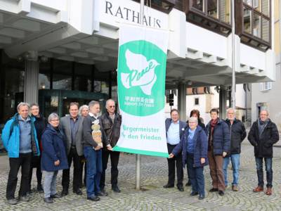 Vor dem Rathaus der Stadt Bretten haben sich mehrere Personen versammelt und nehmen an der Flaggenhissung von Mayors for Peace teil.