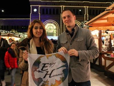 Eine Frau und ein Mann stehen vor der Frontseite des hannoverschen Hauptbahnhofs und halten das Transparent ihrer Communty vor sich. Im Hintergrund ist die Weihnachtsbeleuchtung erkennbar.