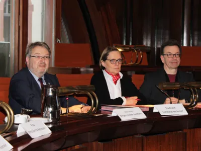 Drei Personen - zwei M&auml;nner und eine Frau - sitzen Im Hodlersaal im Neuen Rathaus hinter Pulten und blicken nach vorne.