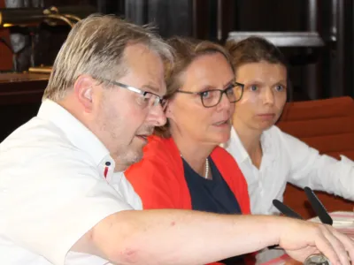Drei Personen - zwei Frauen und ein Mann - sitzen im Hodlersaal hinter Pulten. Die Frau in der Mitte spricht.